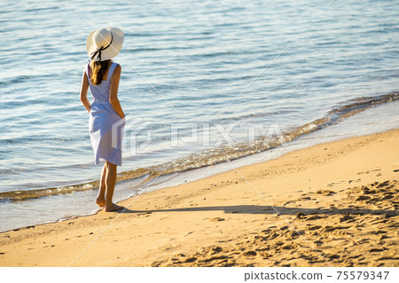 Young woman in straw hat and a dress walking alone on empty sand beach at sea shore. Lonely tourist girl looking at horizon over calm ocean surface on vacation trip. Young woman in straw hat and a dress walking alone on empty sand beach at sea shore. Lonely tourist girl looking at horizon over calm ocean surface on vacation trip. 75579347