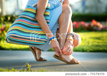 Close up of woman hands tying her open summer sandals shoes on sidewalk in sunny weather. 75579370