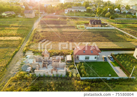 Top down aerial view of two private houses, one under construction with concrete foundament and brick walls and another finished with red tiled roof. 75580804