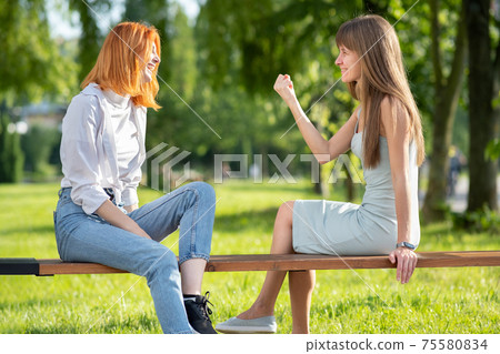 Two young women friends sitting on a bench in summer park and talking having an argument. Two young women friends sitting on a bench in summer park and talking having an argument. 75580834