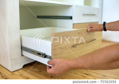 Close up of carpenter hands installing wooden drawer on sliding skids in contemporary cupboard cabinet. 75581059