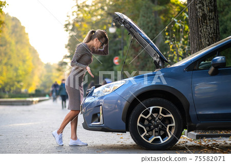 Puzzled female driver standing on a city street near her car with popped up hood looking at broken engine. Puzzled female driver standing on a city street near her car with popped up hood looking at broken engine. 75582071