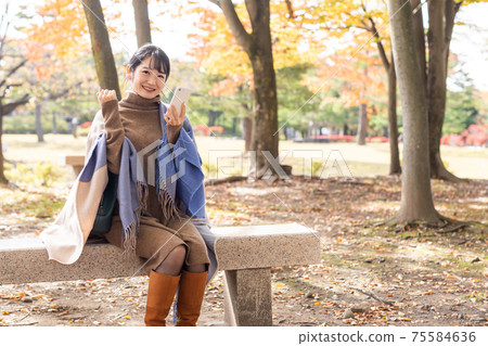 一個女人拿著智能手機，膽量在秋天的公園裡擺姿勢 75584636