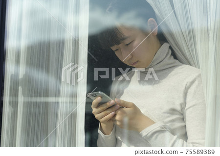 Girl looking at a smartphone by the window (image of request to refrain from going out) 75589389