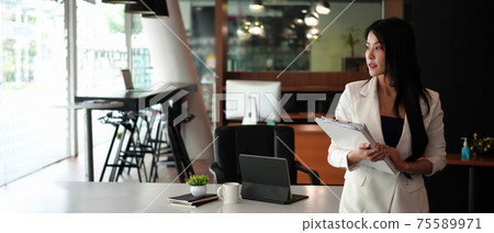 Horizontal photo of confident businesswoman holding files and standing in modern workplace. Horizontal photo of confident businesswoman holding files and standing in modern workplace. 75589971