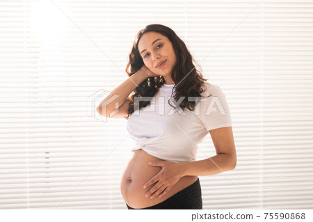 Smiling young beautiful pregnant woman touching her belly and rejoicing. Concept of positive and pleasant feelings while waiting for the baby Smiling young beautiful pregnant woman touching her belly and rejoicing. Concept of positive and pleasant feelings while waiting for the baby 75590868