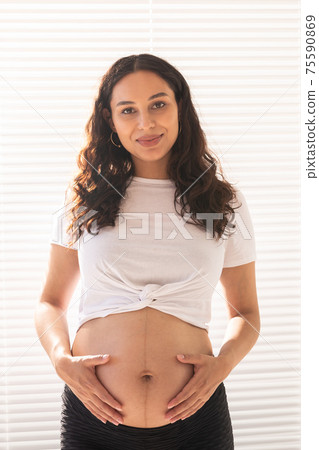 Smiling young beautiful pregnant woman touching her belly and rejoicing. Concept of positive and pleasant feelings while waiting for the baby Smiling young beautiful pregnant woman touching her belly and rejoicing. Concept of positive and pleasant feelings while waiting for the baby 75590869