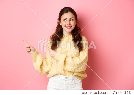 Young happy woman smiling, pointing and looking aside at left copyspace, showing advertisement, standing against pink spring background 75591348