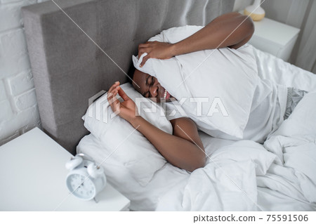 A young man in white tshirt lying in bed and feeling annoyed by the sound of alarm-clock A young man in white tshirt lying in bed and feeling annoyed by the sound of alarm-clock 75591506