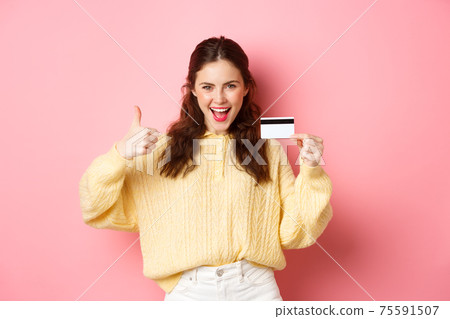 Young modern woman saying yes and showing thumbs up, holding plastic credit card, standing over pink background Young modern woman saying yes and showing thumbs up, holding plastic credit card, standing over pink background 75591507