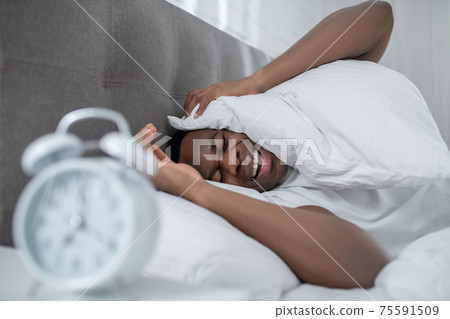 A young man in white tshirt lying in bed and feeling annoyed by the sound of alarm-clock A young man in white tshirt lying in bed and feeling annoyed by the sound of alarm-clock 75591509