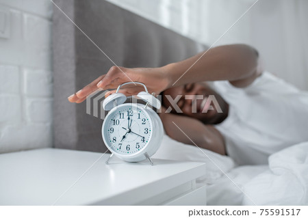 A young man in white tshirt lying in bed and feeling annoyed by the sound of alarm-clock A young man in white tshirt lying in bed and feeling annoyed by the sound of alarm-clock 75591517