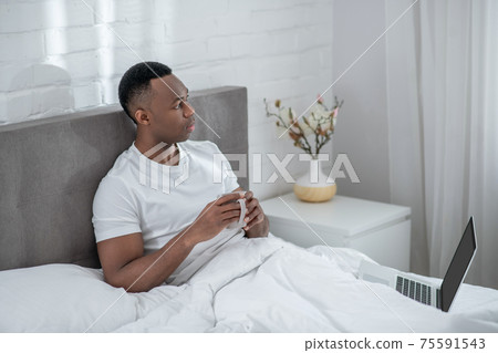 A young man in white tshirt feeling comfortable and having coffee in bed A young man in white tshirt feeling comfortable and having coffee in bed 75591543