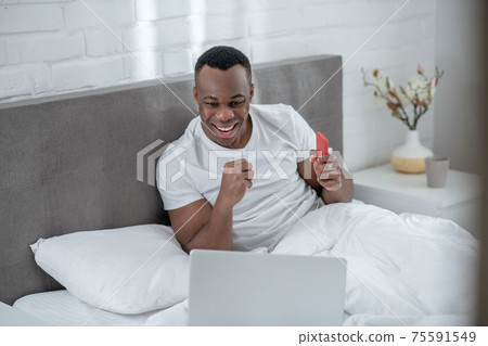 A young man in white tshirt lying in bed and working remotely A young man in white tshirt lying in bed and working remotely 75591549