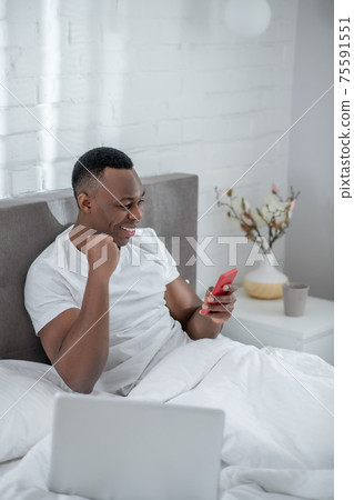 A young man in white tshirt lying in bed and working remotely A young man in white tshirt lying in bed and working remotely 75591551