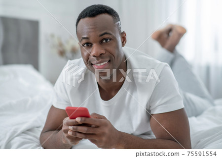 A young man in white tshirt lying in bed and chatting online A young man in white tshirt lying in bed and chatting online 75591554