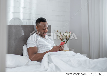 A young man in white tshirt lying in bed and watching something on his smartphone A young man in white tshirt lying in bed and watching something on his smartphone 75591630