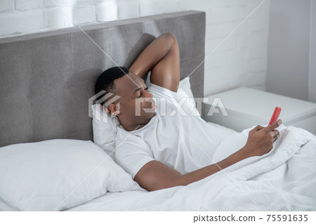 A young man in white tshirt lying in bed and watching something on his smartphone A young man in white tshirt lying in bed and watching something on his smartphone 75591635