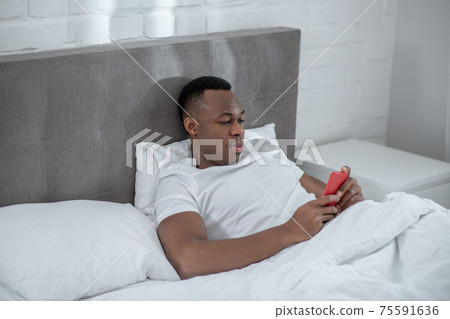 A young man in white tshirt lying in bed and watching something on his smartphone A young man in white tshirt lying in bed and watching something on his smartphone 75591636