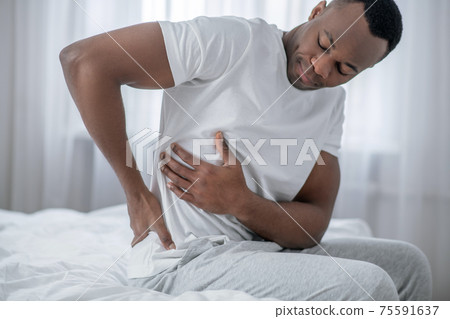 African american man sitting on bed and having lower bakc pain African american man sitting on bed and having lower bakc pain 75591637