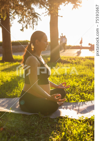 Cute young caucasian pregnant woman is meditating while sitting on a rug on the lawn on a sunny summer evening. Concept of pacification and energy boost 75591667
