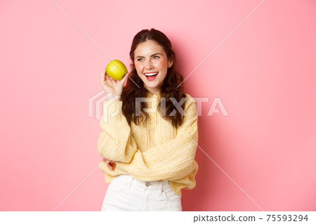 Healthcare and lifestyle concept. Cheerful smiling woman, showing her white perfect smile and green apple near face, eating healthy fruits, standing against pink background Healthcare and lifestyle concept. Cheerful smiling woman, showing her white perfect smile and green apple near face, eating healthy fruits, standing against pink background 75593294