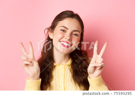 Close up of beautiful young woman showing peace v-sign and smiling happy at camera, wearing bright glam make up, standing against pink background 75593295