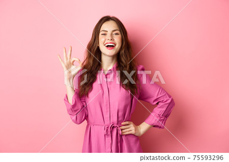 Young attractive woman saying yes, showing okay sign in approval, smiling confident and satisfied, standing over pink background Young attractive woman saying yes, showing okay sign in approval, smiling confident and satisfied, standing over pink background 75593296