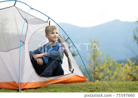 Happy child boy resting in a tourist tent at mountain campsite enjoying view of beautiful summer nature. Hiking and active way of life concept. Happy child boy resting in a tourist tent at mountain campsite enjoying view of beautiful summer nature. Hiking and active way of life concept. 75597660