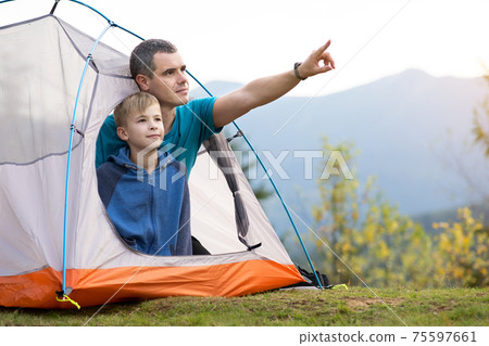Young father and his child son hiking together in summer mountains. Active family resting in tourist tent. Young father and his child son hiking together in summer mountains. Active family resting in tourist tent. 75597661