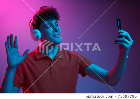 Young man in headphones with phone isolated on gradient background in neon light 75597790
