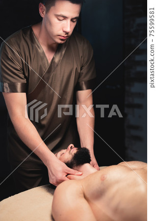 Professional neck massage to a bearded male athlete in a dark room of a spa massage room Professional neck massage to a bearded male athlete in a dark room of a spa massage room 75599581