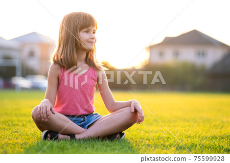 Young pretty child girl sitting on fresh green grass lawn on warm summer day outdoors. 75599928