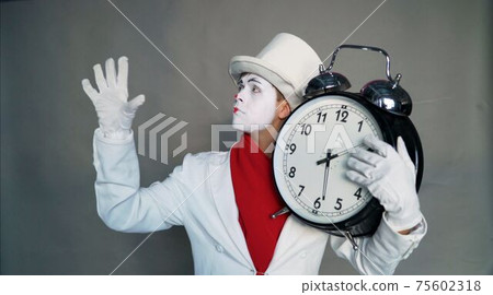 A man in a clown suit stands beside men in business suits, who sit at the desk. He holds a big clock in his hand. They are in a bright big office. Clown with a big clock in his hands A man in a clown suit stands beside men in business suits, who sit at the desk. He holds a big clock in his hand. They are in a bright big office. Clown with a big clock in his hands 75602318