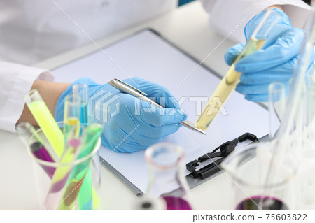 Scientist chemist holding test tube with yellow liquid in his hands and writing in documents closeup 75603822