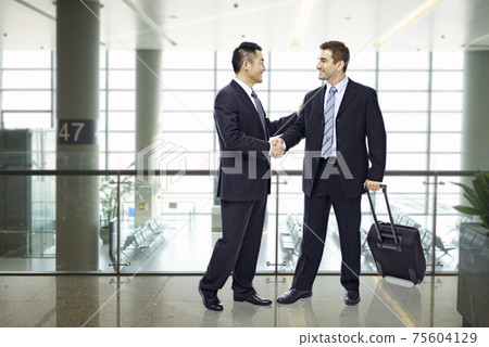 two businessmen, one asian and one caucasian, shaking hands and smiling at modern airport 75604129
