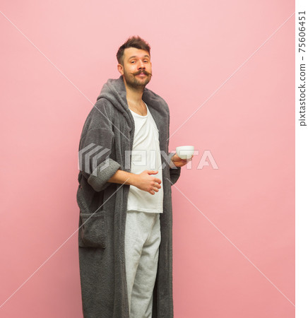 Pleased man in gray bathrobe with cup of coffee isolated on pink background 75606451