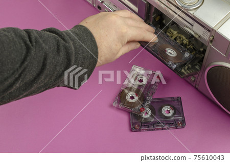 Hand inserts the audio cassette into the boom box. Boom box Stereo Radio Cassette Recorder and random cassettes on pink background. Vintage Cassette Deck, retro technology, selective focus. Hand inserts the audio cassette into the boom box. Boom box Stereo Radio Cassette Recorder and random cassettes on pink background. Vintage Cassette Deck, retro technology, selective focus. 75610043