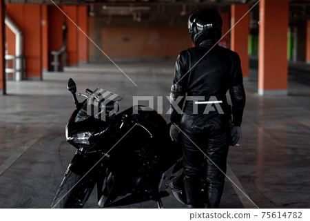 Stylish young woman motorcycle rider in black protective gear and full-face helmet near her bike on underground parking. 75614782