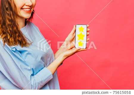 Stylish caucasian girl in a pale blue t-shirt on a pink background. Hand holds a smartphone with a star icon as a top-ranking concept. Stylish caucasian girl in a pale blue t-shirt on a pink background. Hand holds a smartphone with a star icon as a top-ranking concept. 75615058