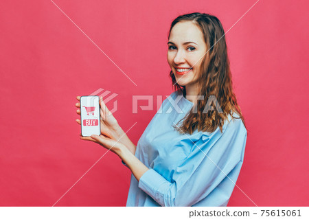 Stylish caucasian girl in a pale blue t-shirt on a pink background. Online shopping concept - button - buy next to an abstract shopping cart in a smartphone. Stylish caucasian girl in a pale blue t-shirt on a pink background. Online shopping concept - button - buy next to an abstract shopping cart in a smartphone. 75615061