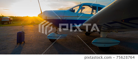A blue suitcase and a parked small private jet. Quadruple plane with a propeller for an air taxi in the sunset. Self travel concept. plane for VIP persons. 75628764
