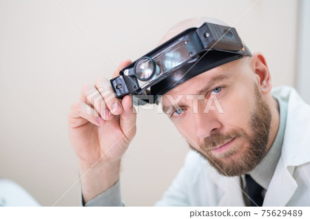 Male bearded doctor in a medical coat and with a magnifying head strap w lights sitting on the desk. Optometrist equipment. Medical office. Male bearded doctor in a medical coat and with a magnifying head strap w lights sitting on the desk. Optometrist equipment. Medical office. 75629489