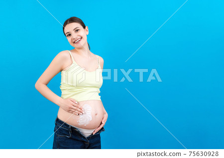 pregnant woman in unzipped jeans applying moisturizing cream on her belly against stretch marks at colorful background with copy space. Skin care concept 75630928