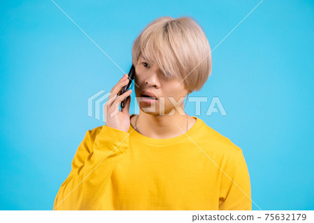 Young surprised asian man speaks with smile on phone. Guy with dyed blond hair holding and using 75632179