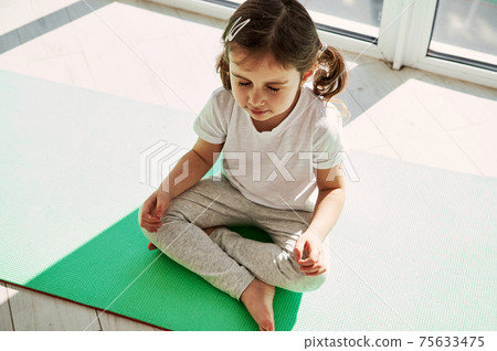 Adorable girl with ponytails is sitting on an exercise mat and doing meditation. Developing healthy habits from early childhood. 75633475