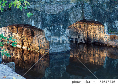 Old, small stone bridge close-up across small river, warm sunlight reflected from arches bridge, calm water surface 75637136