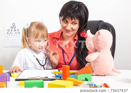 A woman doctor gave a stethoscope to a little caucasian girl 3-4 years old in an office to listen to. Children s doctor s office with toys, children s medicine 75637437