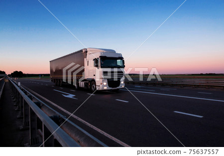 A white semi-trailer truck drives down the highway in the evening with its headlights on. Blue sky and sunset. The mode of work and rest of truck drivers. Copy space for text A white semi-trailer truck drives down the highway in the evening with its headlights on. Blue sky and sunset. The mode of work and rest of truck drivers. Copy space for text 75637557