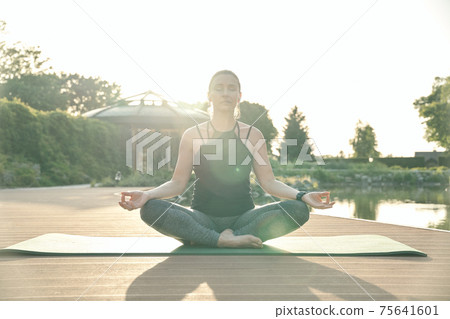 Quiet mind. Relaxed middle aged woman meditating, sitting in Lotus pose while doing yoga on a mat in a green park Quiet mind. Relaxed middle aged woman meditating, sitting in Lotus pose while doing yoga on a mat in a green park 75641601
